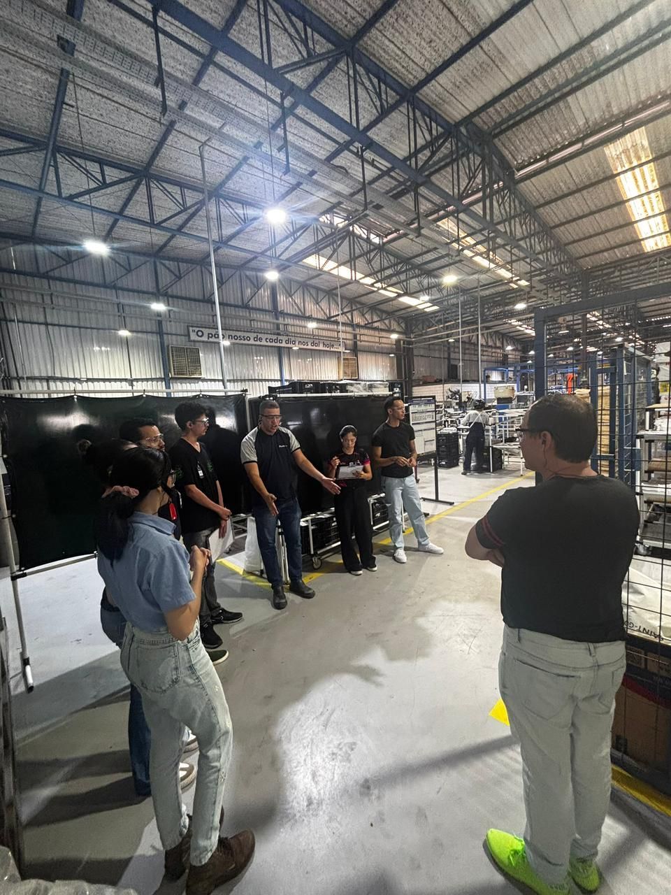 Pessoas em um ambiente fabril, algumas ouvindo um homem falar. Galpão cinza com pé-direito alto.