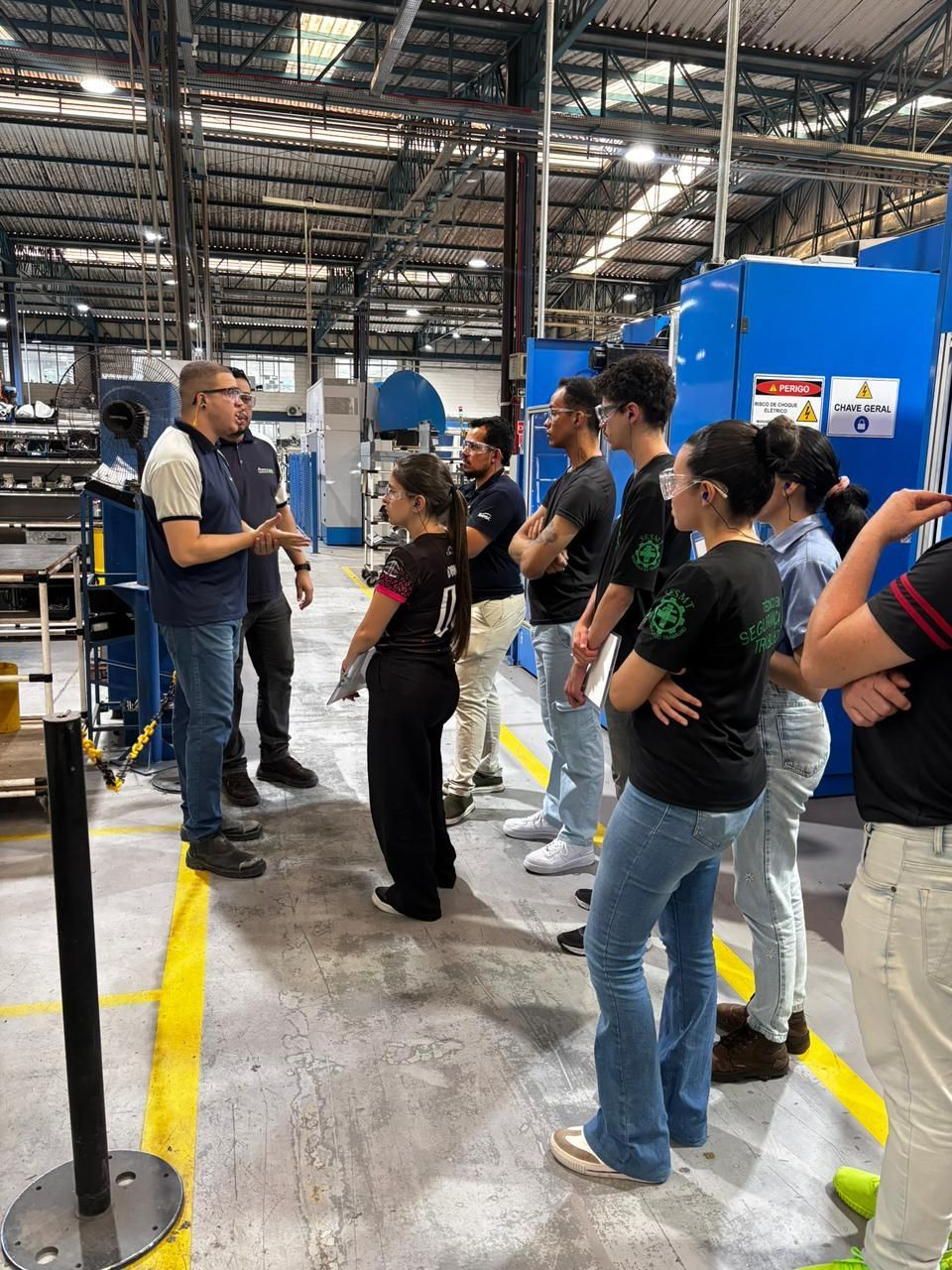 Grupo de pessoas em visita guiada a uma fábrica; o guia fala, enquanto os outros ouvem perto das máquinas.