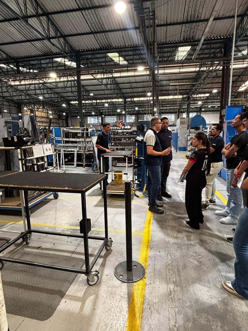 Grupo de pessoas em uma fábrica, ouvindo um homem falar. Ambiente industrial com máquinas e bancadas de trabalho.