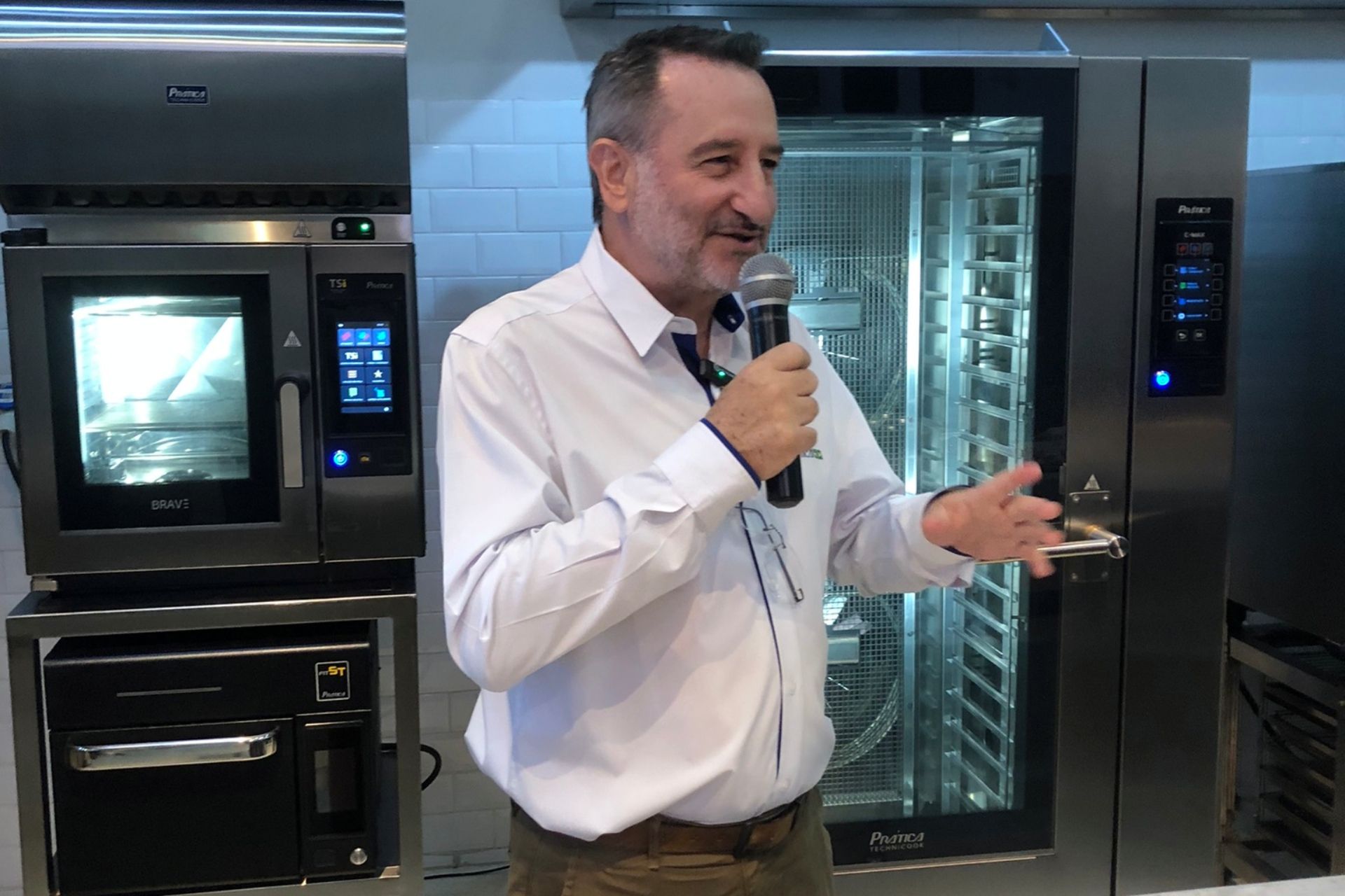 Homem falando ao microfone, em pé perto de fornos industriais em uma cozinha.
