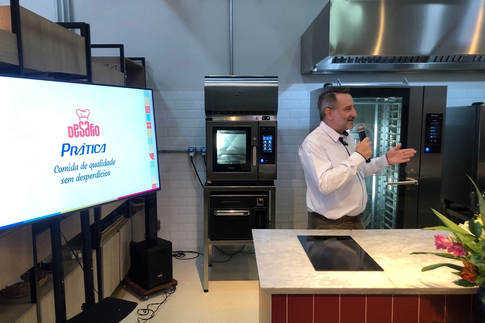 Homem falando durante uma apresentação de comida; forno e tela exibindo informações.