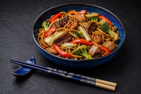 Tigela azul de macarrão frito com legumes e pauzinhos sobre uma mesa escura.