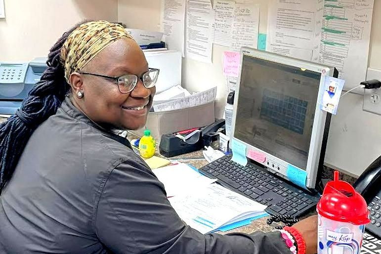 Woman at computer smiling, in an office setting. Wearing glasses and a headscarf, looking towards the camera in Fayetteville, NC.