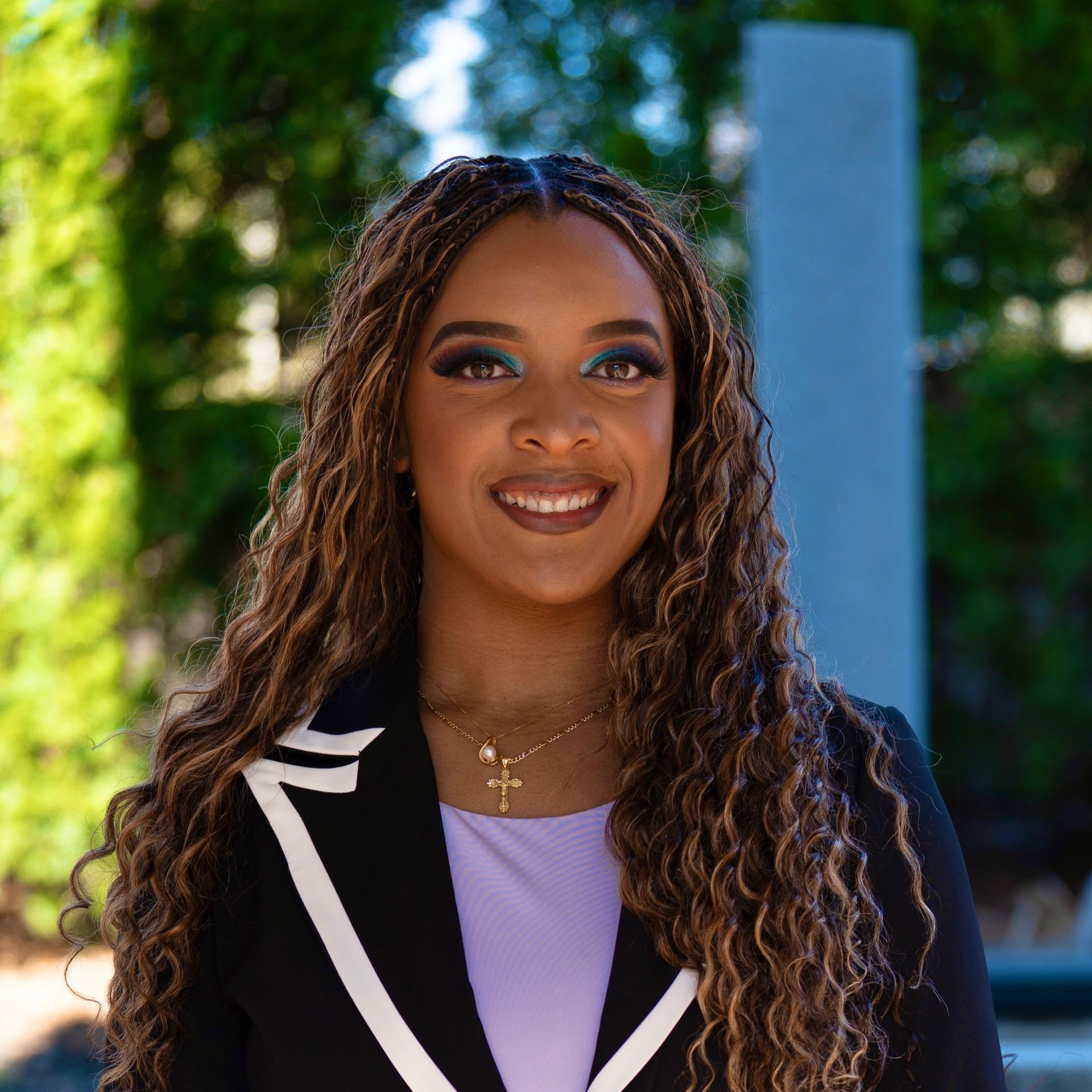 Smiling woman in black blazer and lavender top outdoors with blurred greenery and blue column background