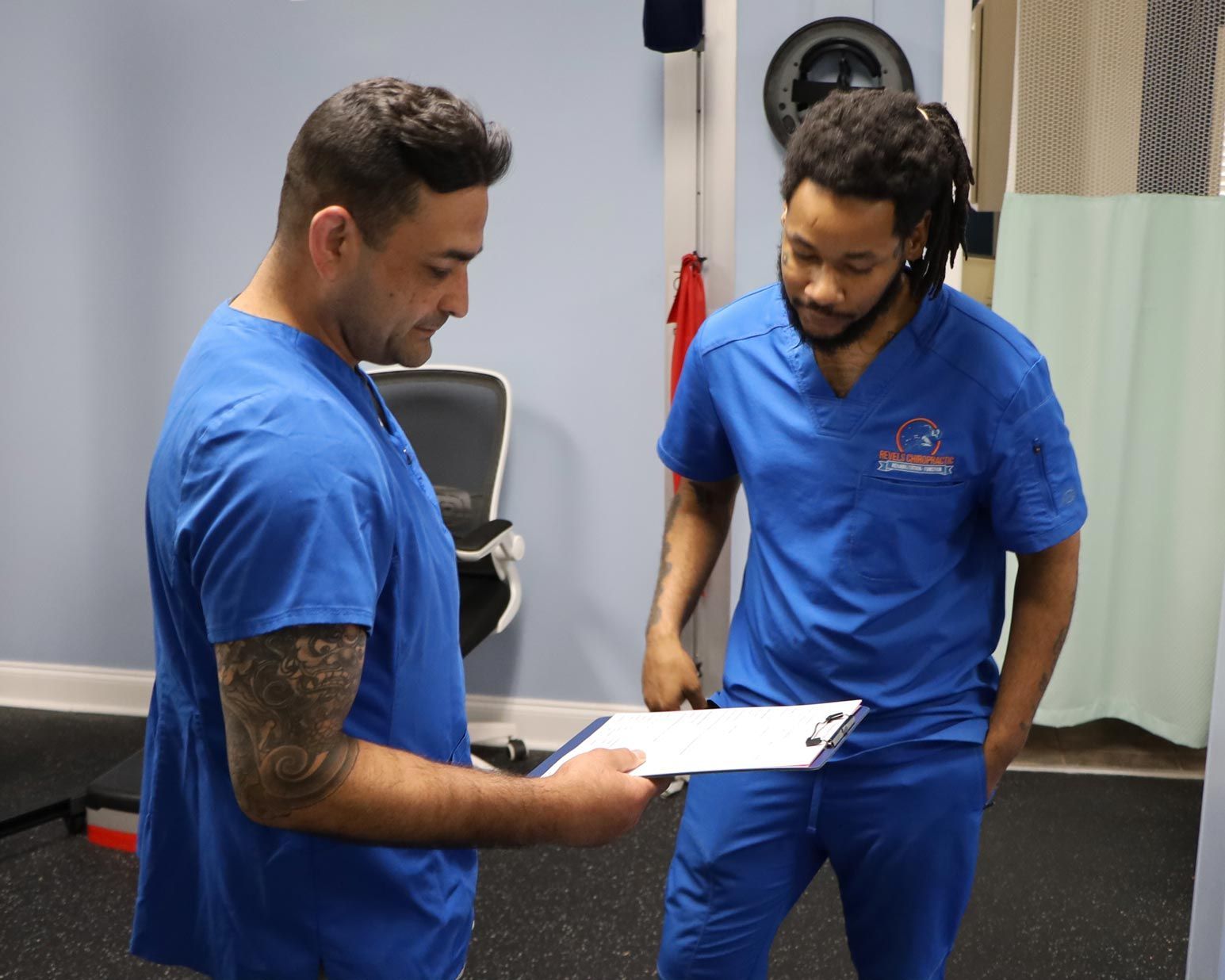 Two men in blue scrubs are standing next to each other looking at a clipboard.