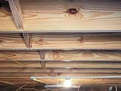 Wooden joists and plywood subfloor in a crawl space, with pipes and wiring visible below.
