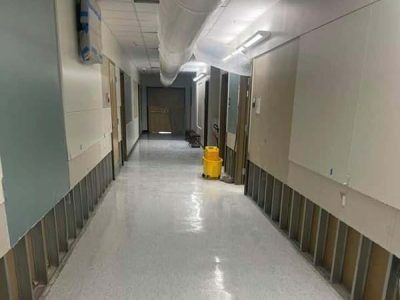 Construction in a school hallway: exposed wall frames, white floor, and a yellow bucket.