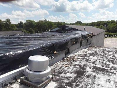 Roof of a building with a large black tarp covering a portion; a ventilation unit is in the foreground.