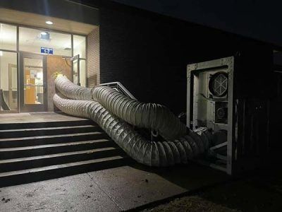 Two large ventilation tubes snake from a door of a building to an outside unit at night.