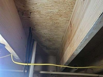 Wooden beams and OSB subfloor in an unfinished basement, with electrical wiring and plumbing visible.