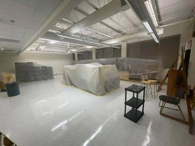 Large, empty art studio with white floor and ceiling. Covered objects, table, stools, and storage shelves present.
