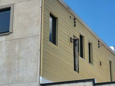 Exterior of a building with light yellow siding and a concrete wall. Three windows are visible.