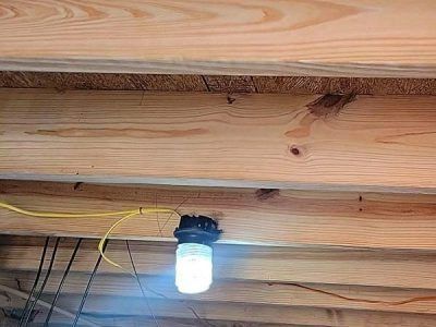 A light bulb in a black socket hangs from wood beams in a dim setting. Yellow wire attached.