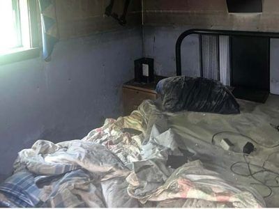 Bedroom scene: messy bed with tangled sheets, nightstand, window, dark stains on wall.