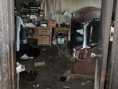 Room interior, possibly damaged by fire. Debris covers the floor, furniture charred, door frame visible.