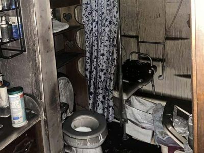 A bathroom heavily damaged by fire; soot covers walls, toilet, and shelving; shower curtain hangs.
