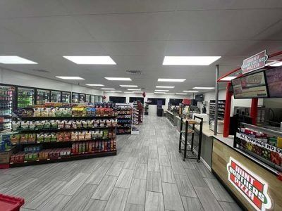 Interior of a convenience store with shelves of snacks and drinks, counter, and coolers. Gray and red colors.