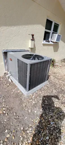 An air conditioner outside a building. It's gray with a black fan and is on a concrete base. A window is above it.