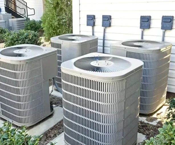 Four gray air conditioning units outside, with electrical outlets above them and some greenery around.