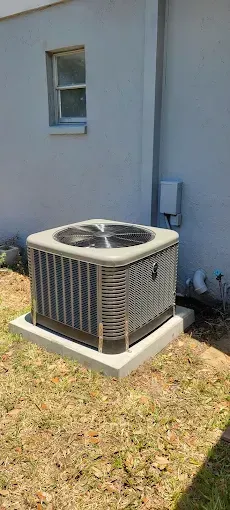 An air conditioning unit sits on a concrete pad next to a light gray building and grass.