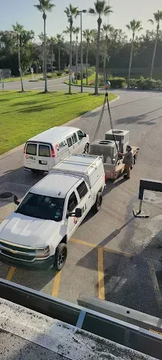 White trucks and van near a grassy area with palm trees. A trailer is holding large equipment, possibly HVAC units.
