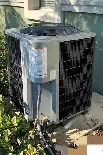 Outdoor air conditioning unit next to bushes. Gray metal with black grill and white information label.