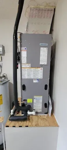 HVAC unit on a wooden platform against a white wall; black pipe; water heater to the left.
