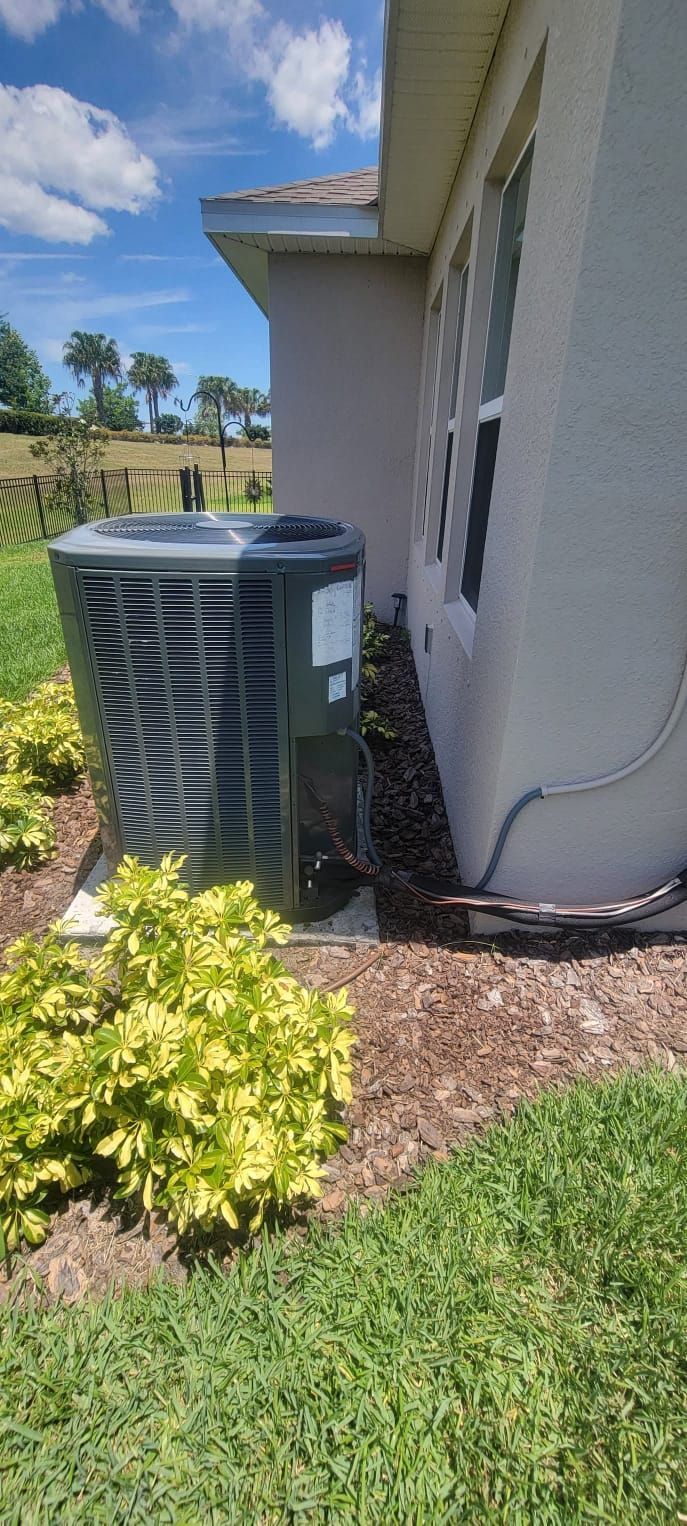 An air conditioning unit next to a house with mulch and a small shrub. Blue sky in the background.