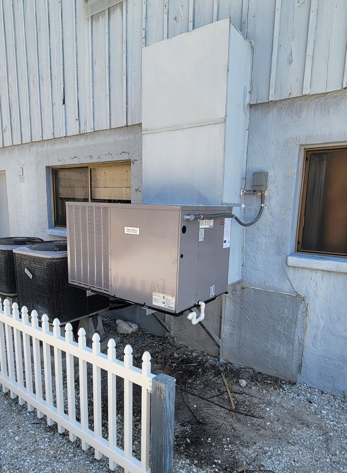 An outdoor HVAC unit attached to a white building with a rectangular metal duct.