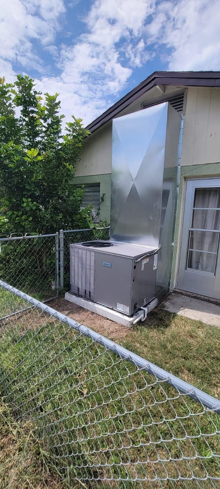 An air conditioning unit with a large metal duct next to a chain-link fence, under a cloudy sky.