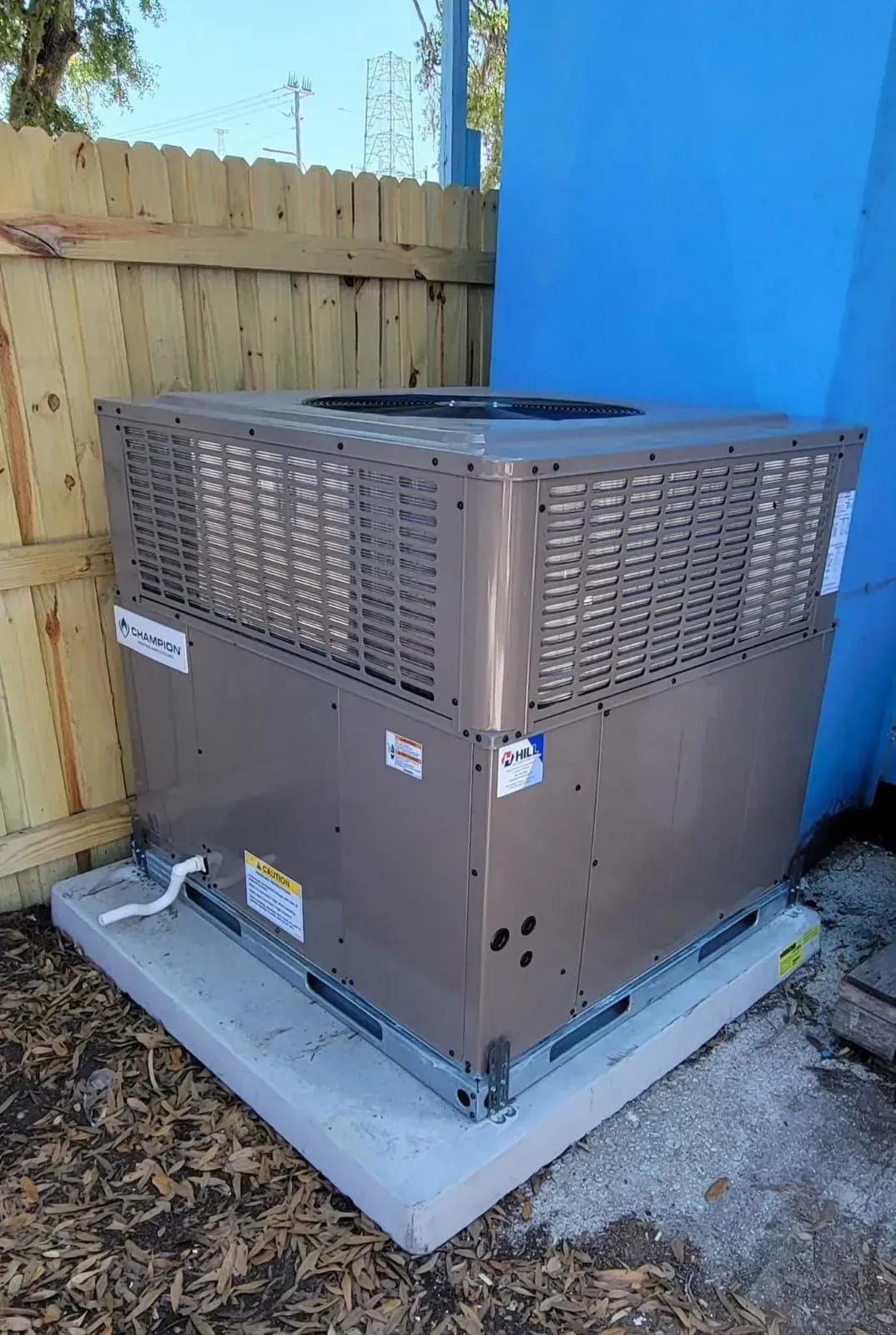 Large, beige air conditioning unit on a concrete pad next to a blue wall and a wooden fence.