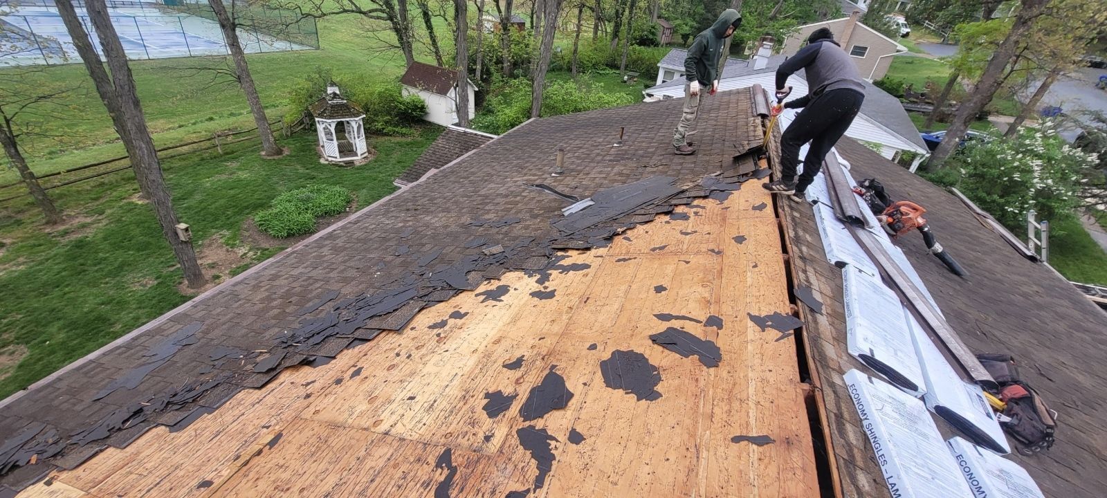 Worker repairing a roof with exposed plywood and roofing materials on a house surrounded by trees