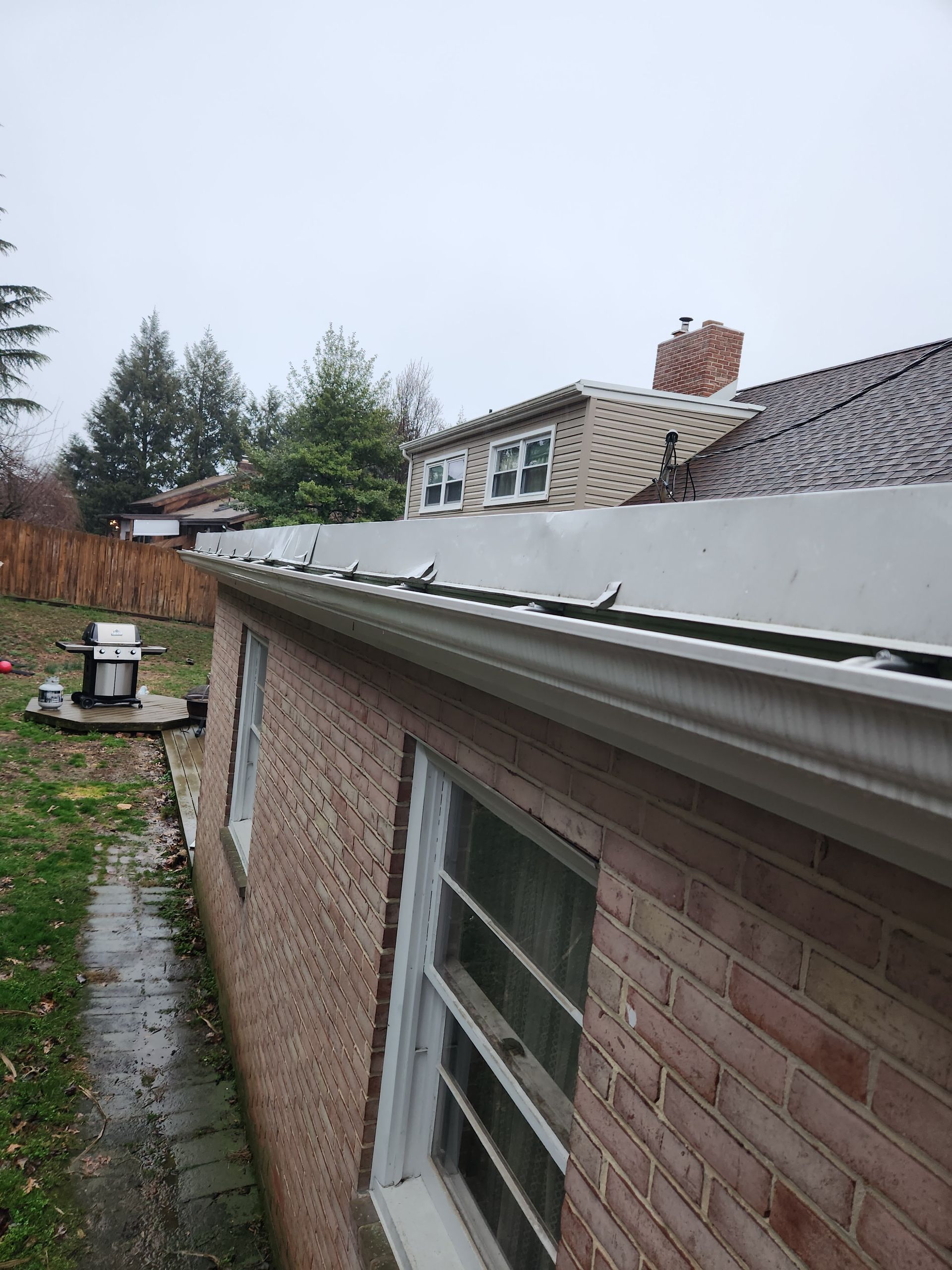A view of a brick house exterior featuring a white gutter system and a roof with light-colored shingles.