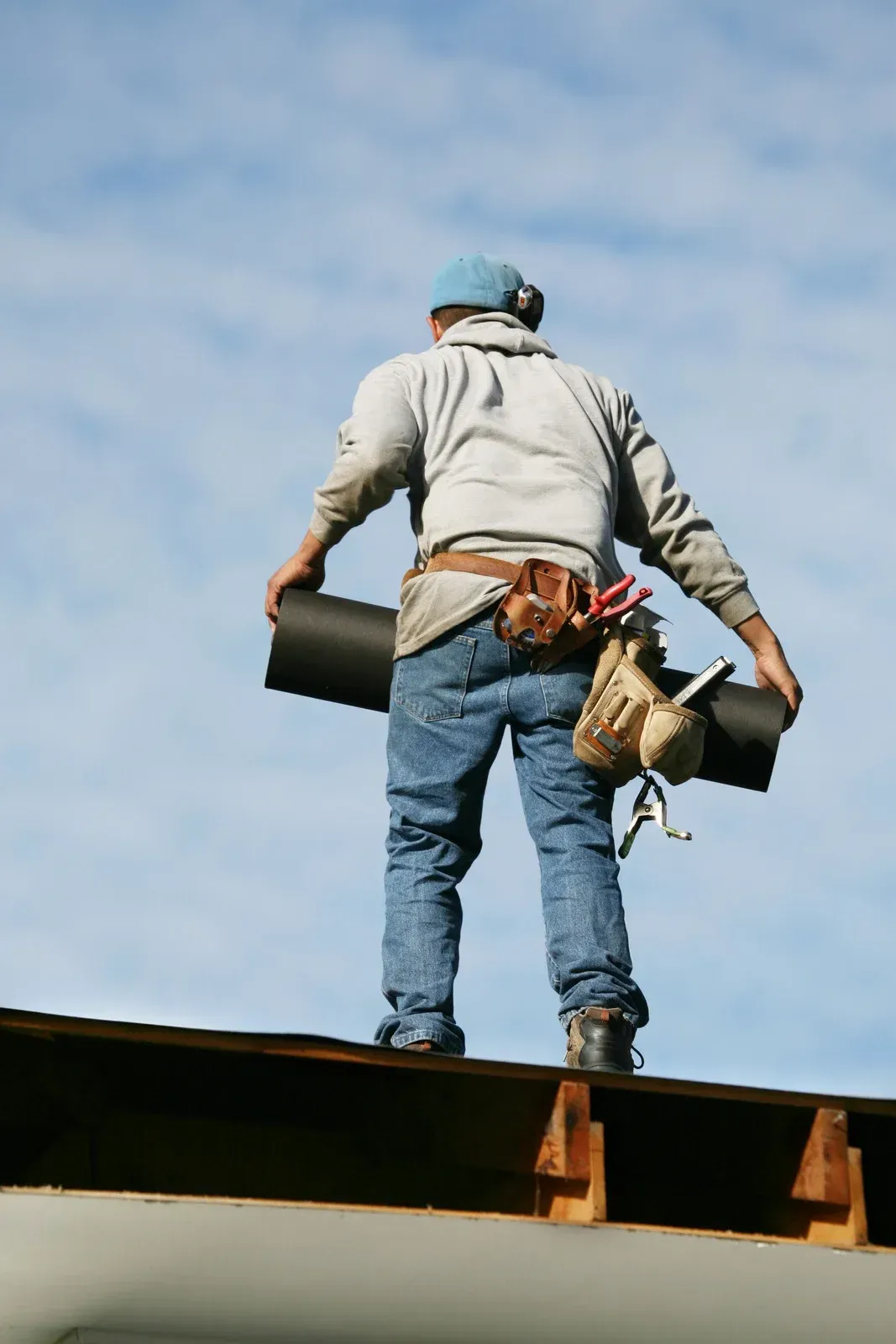 Roofer on a roof holding rolled roofing material against a cloudy sky.