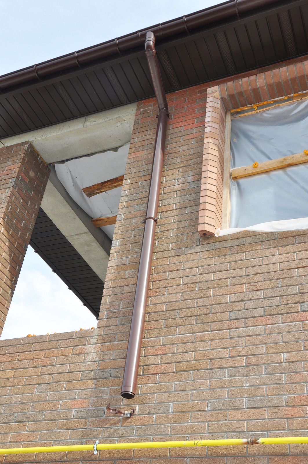 Brown rain gutter on a brick building with a window covered in plastic.