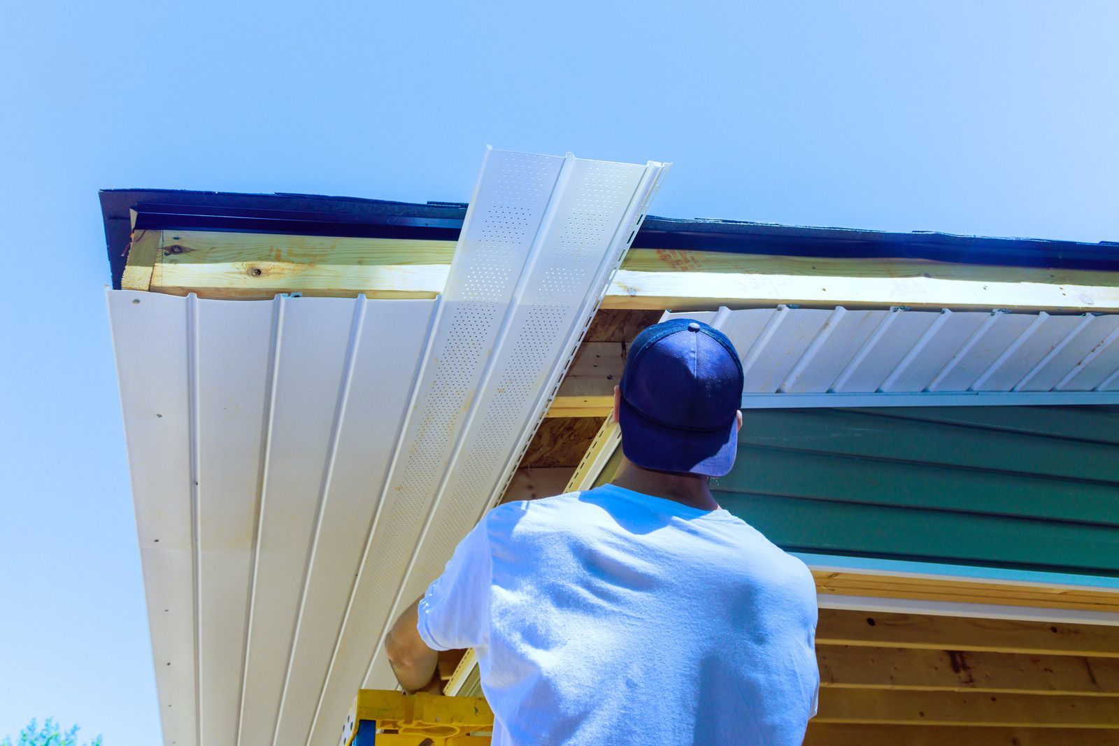 Person installing white siding on a building's roof under a blue sky.