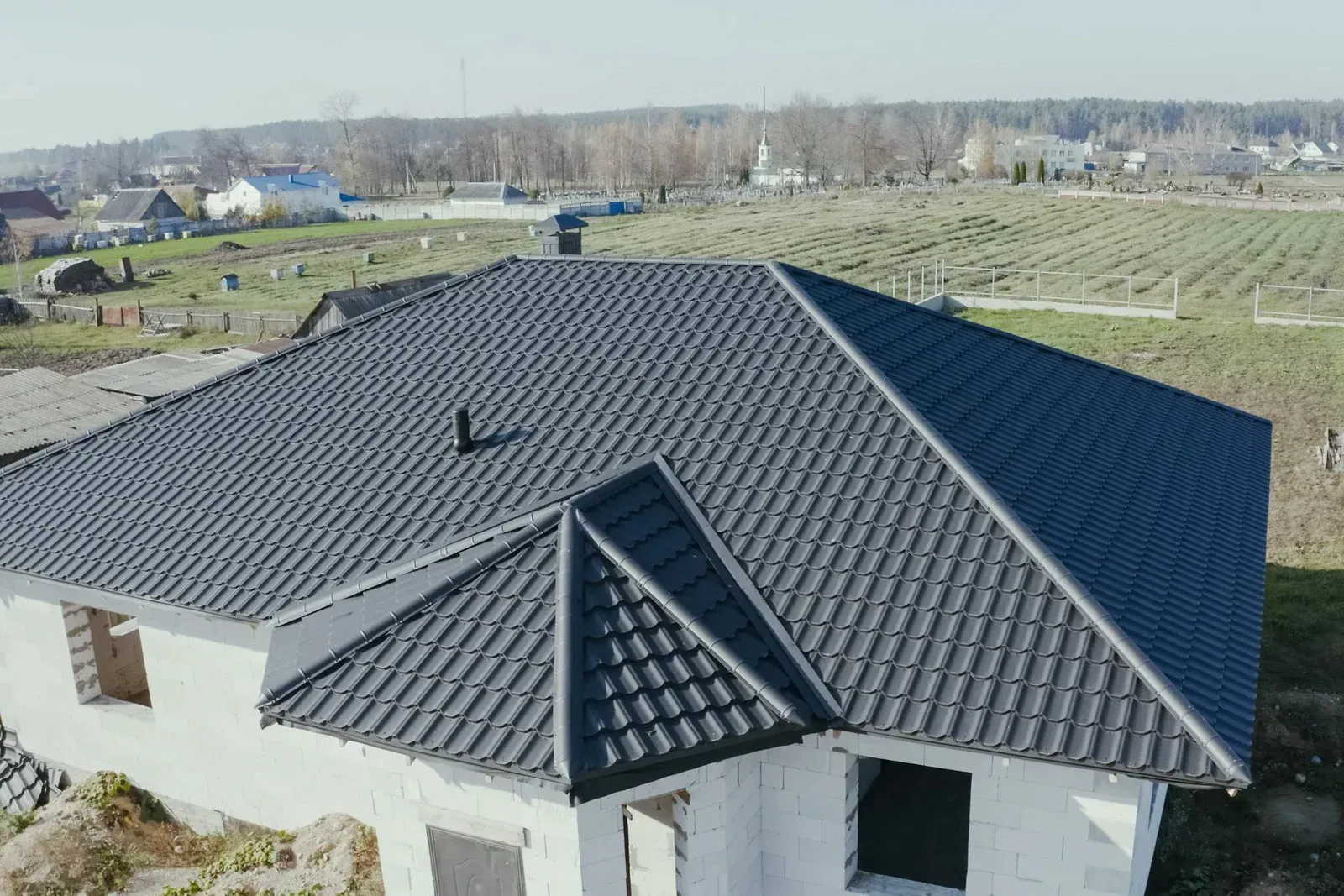 Gray tiled roof on a partially constructed white house; fields and trees in background.