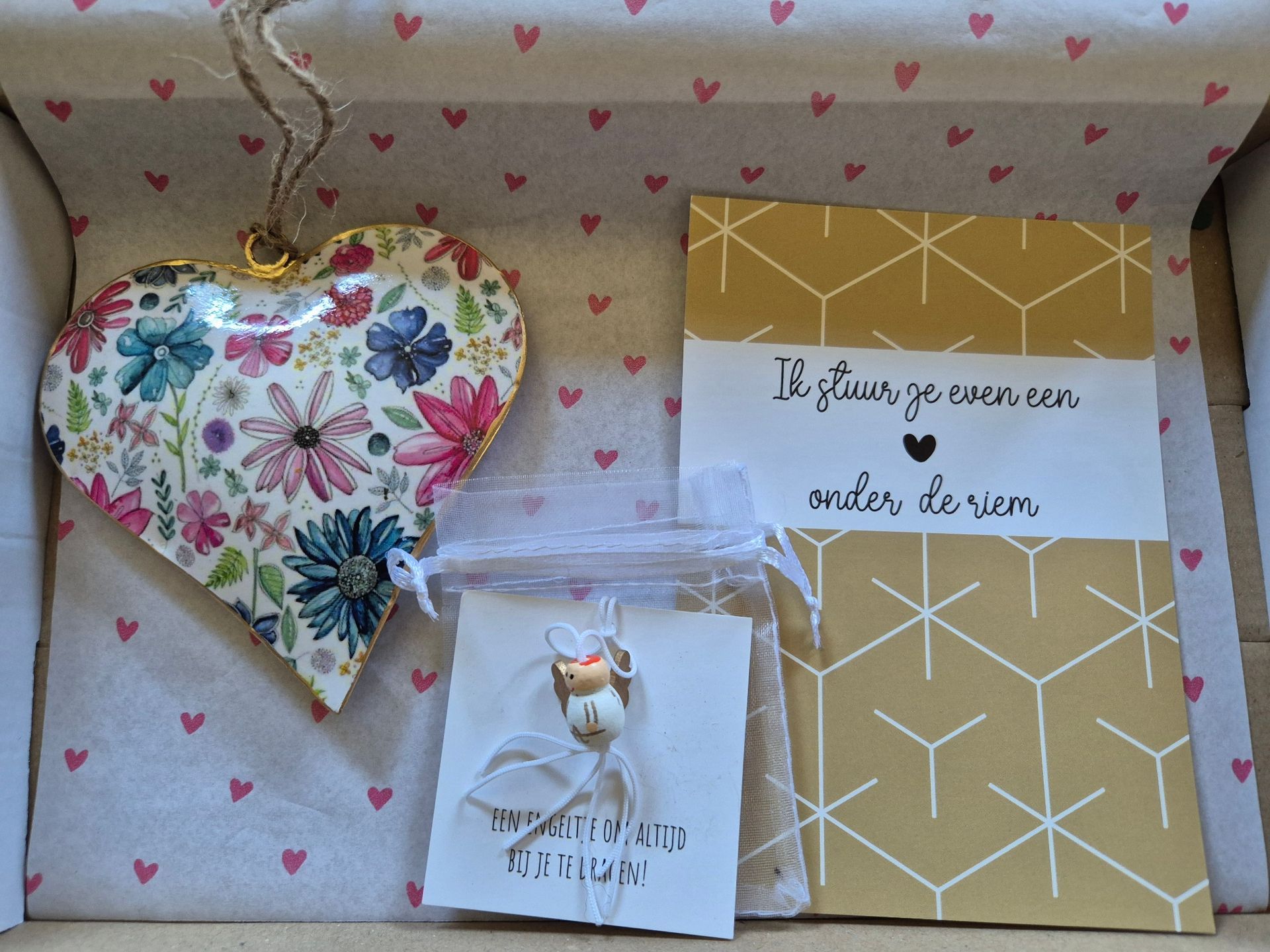 Decoratie met een hartvormig bloemenmotief, een cadeautasje met een bedeltje en een kaartje met een goudkleurig geometrisch patroon en witte tekst op een houten ondergrond.
