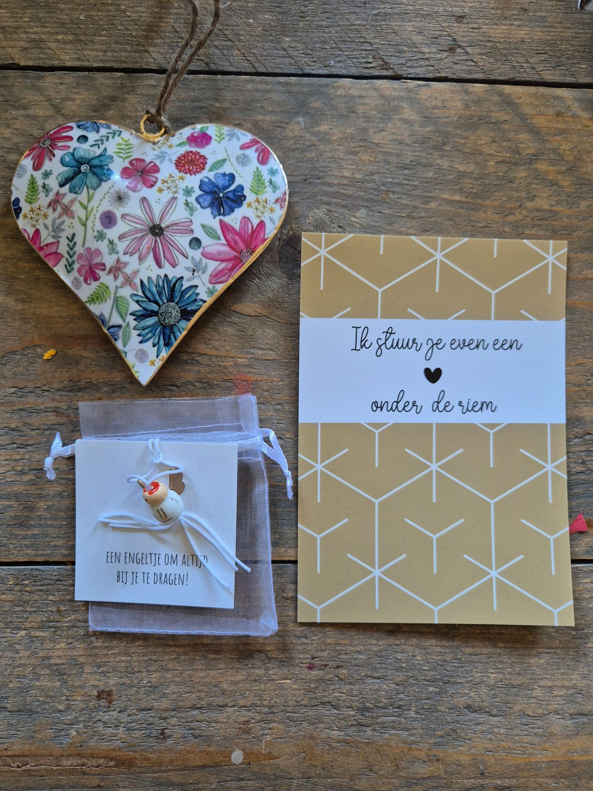 Decoratie met een hartvormig bloemenmotief, een cadeautasje met een bedeltje en een kaartje met een goudkleurig geometrisch patroon en witte tekst op een houten ondergrond.