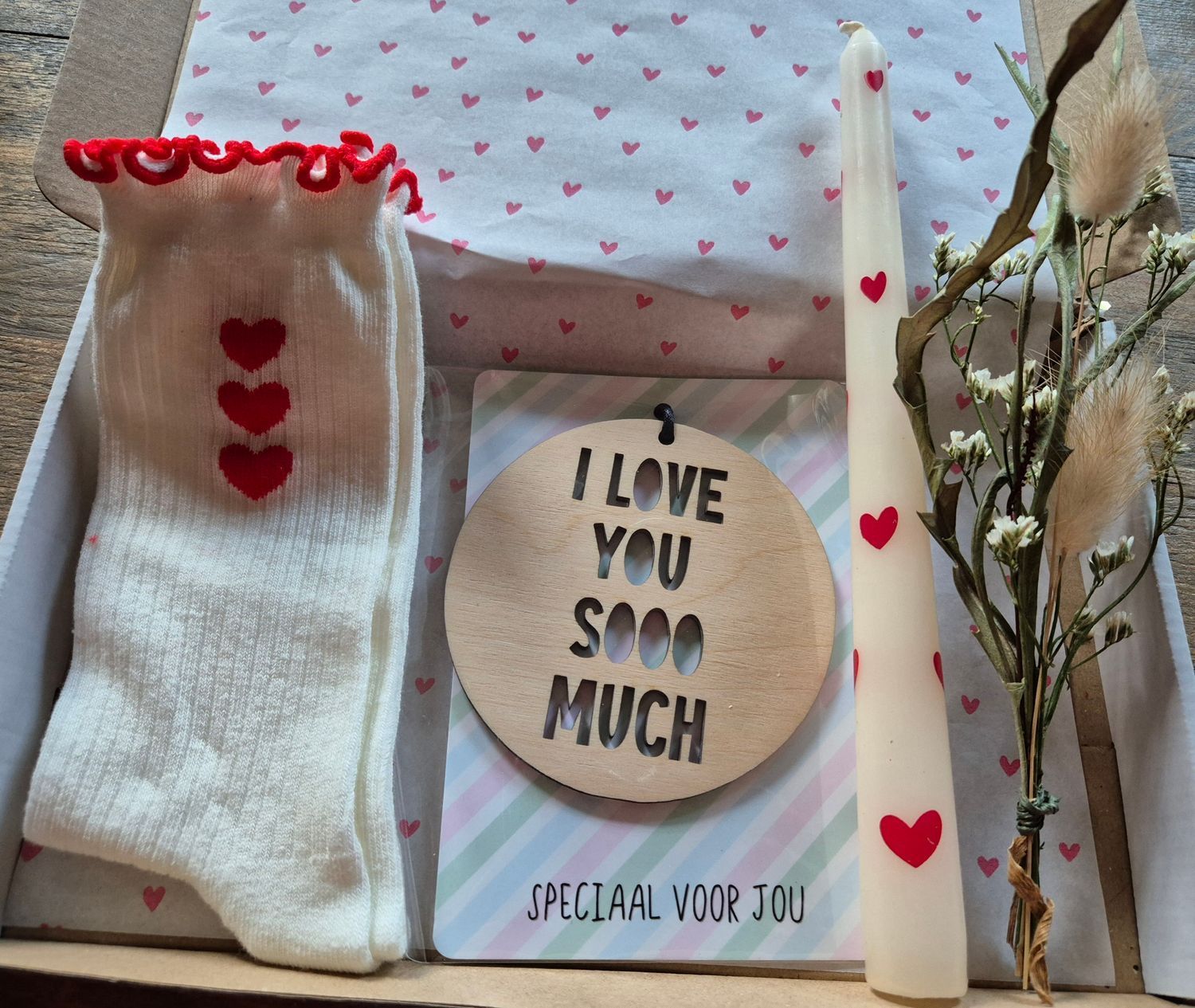 Cadeauverpakking met sokken met hartjesmotief, een kaars, een houten bordje en bloemen. In witte en rode kleuren.