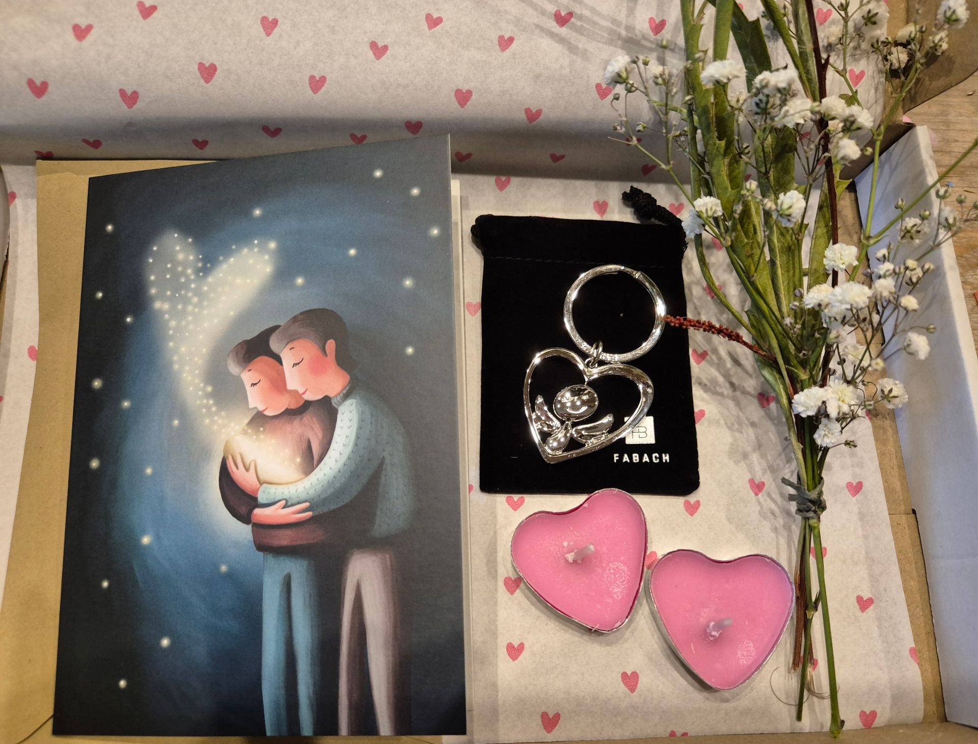 Cadeauverpakking met een kaartje, sleutelhangertje, hartvormige kaarsen en bloemen. Op het kaartje staan ​​twee mensen die elkaar omarmen met een stralend hart.