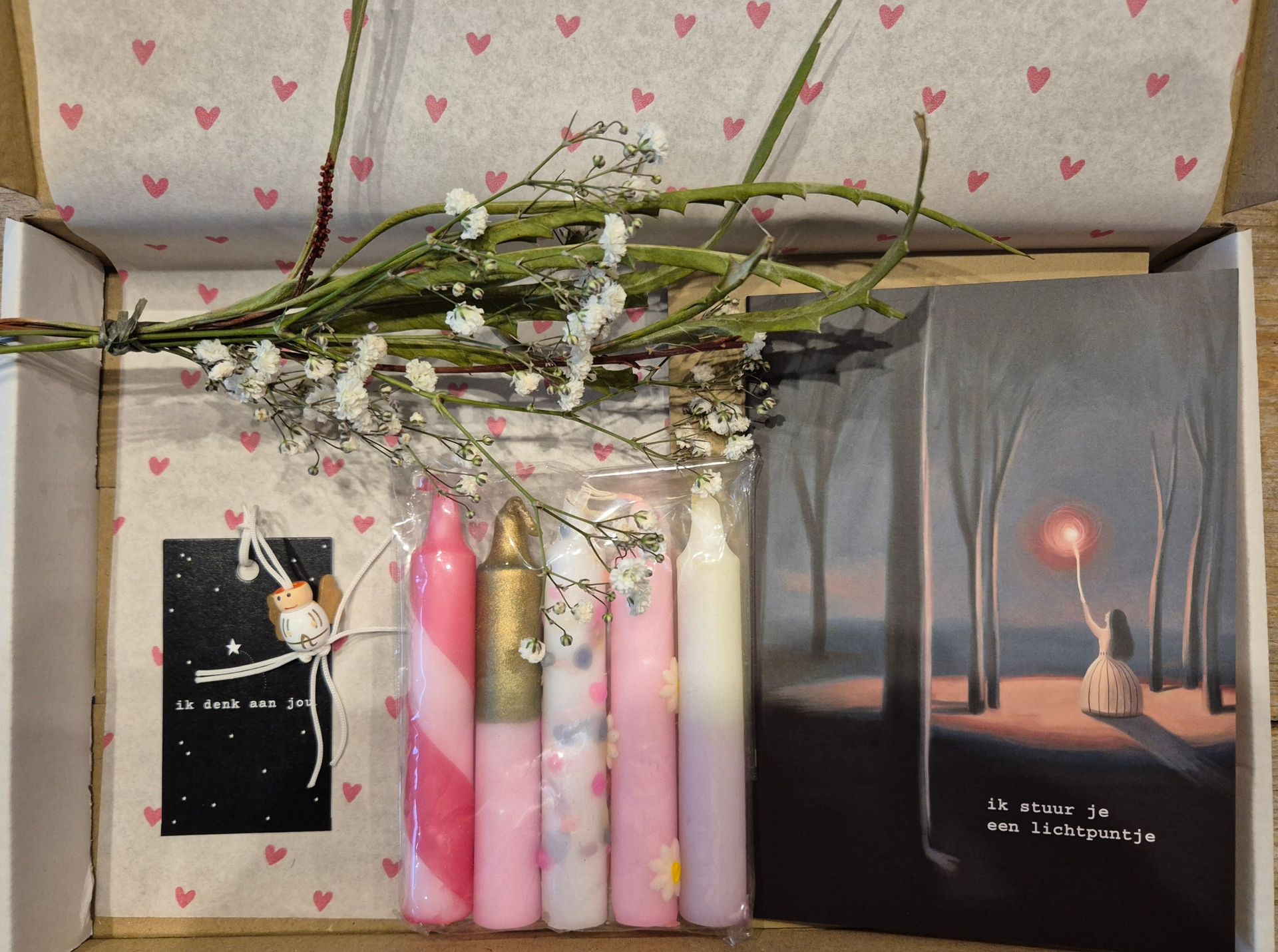 Een geschenkdoos met kaarsen, bloemen en een kaart met een schilderij erop. Roze en witte tinten.