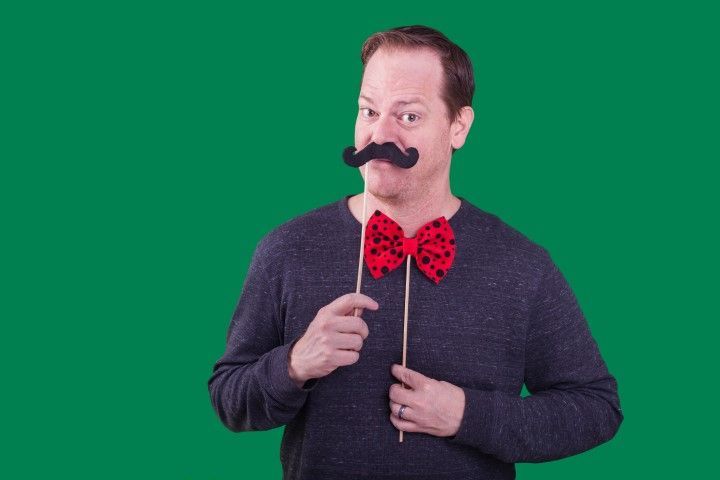 Man holding a fake mustache and wand, wearing a red bow tie against a green background