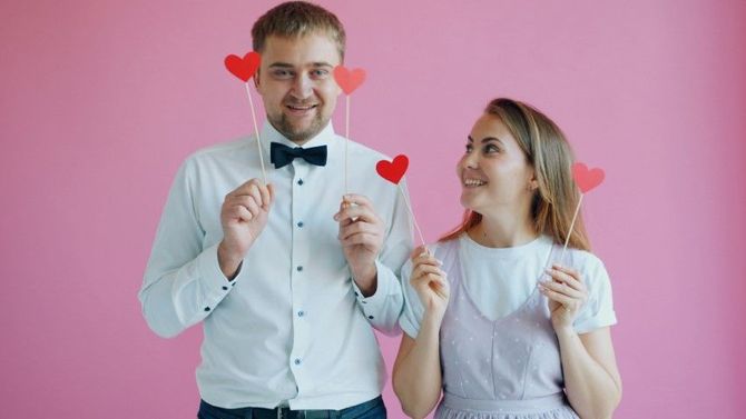 Two people holding heart-shaped signs against a pink background, smiling at each other.