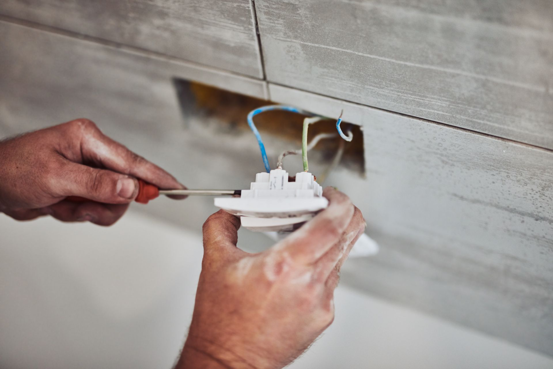 Hands installing a socket outlet into a tiled wall, holding a screwdriver and the outlet.