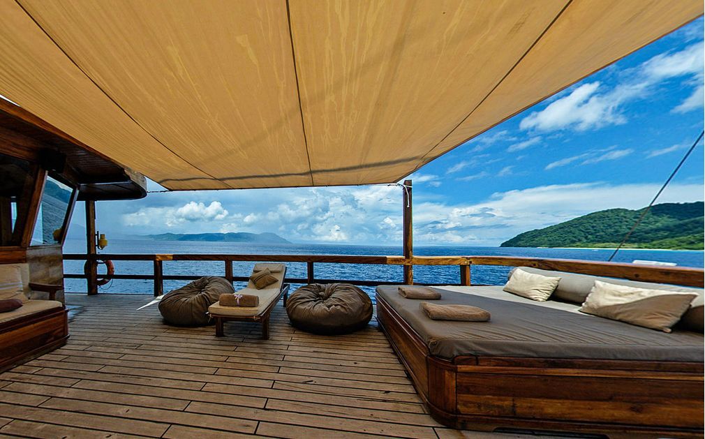 Deck of a boat with cushioned lounge and bean bag chairs under a sunshade, overlooking ocean and distant islands.