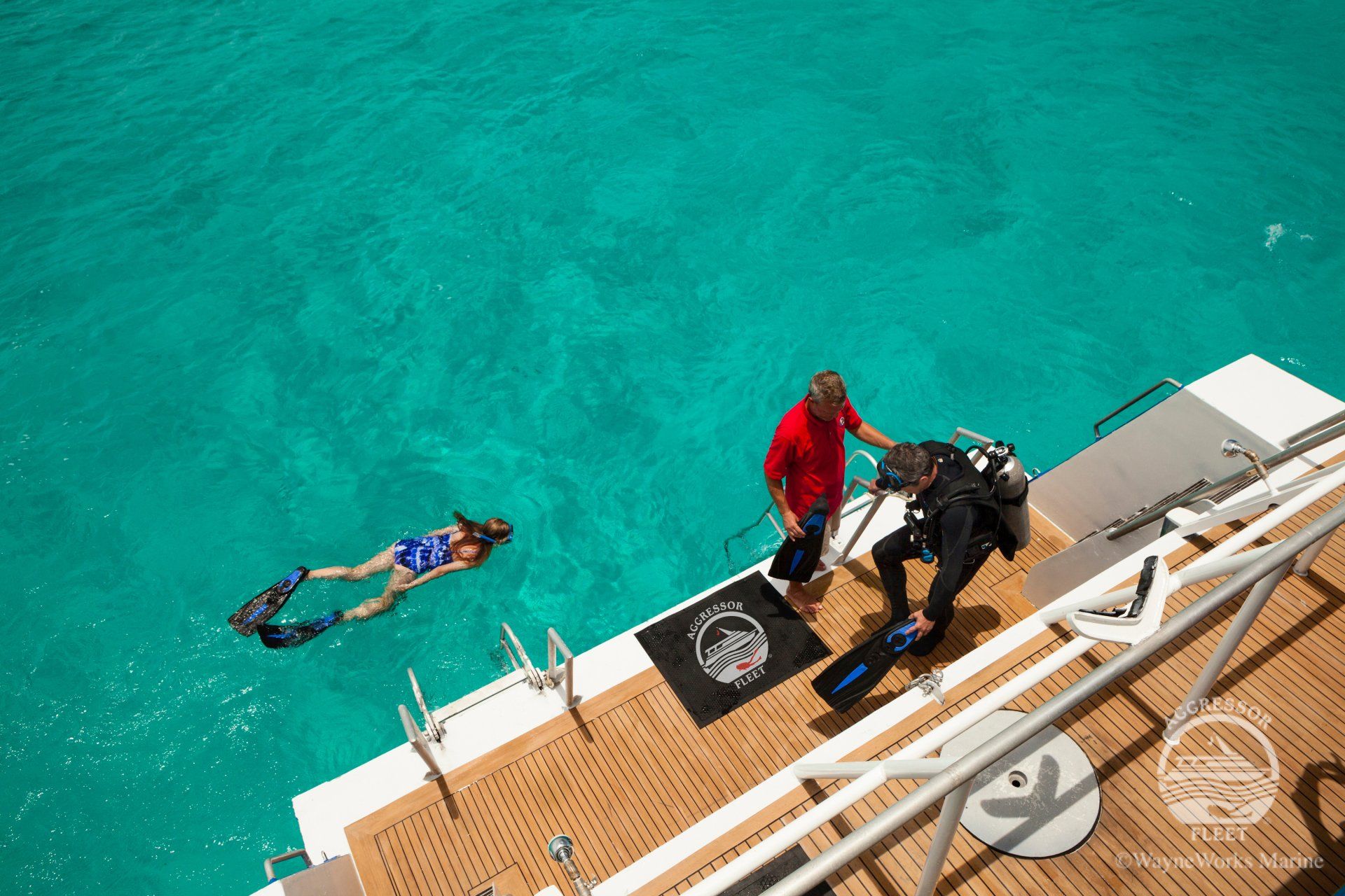 Divers preparing to enter turquoise water from a boat. One swims, one dons gear, another assists.