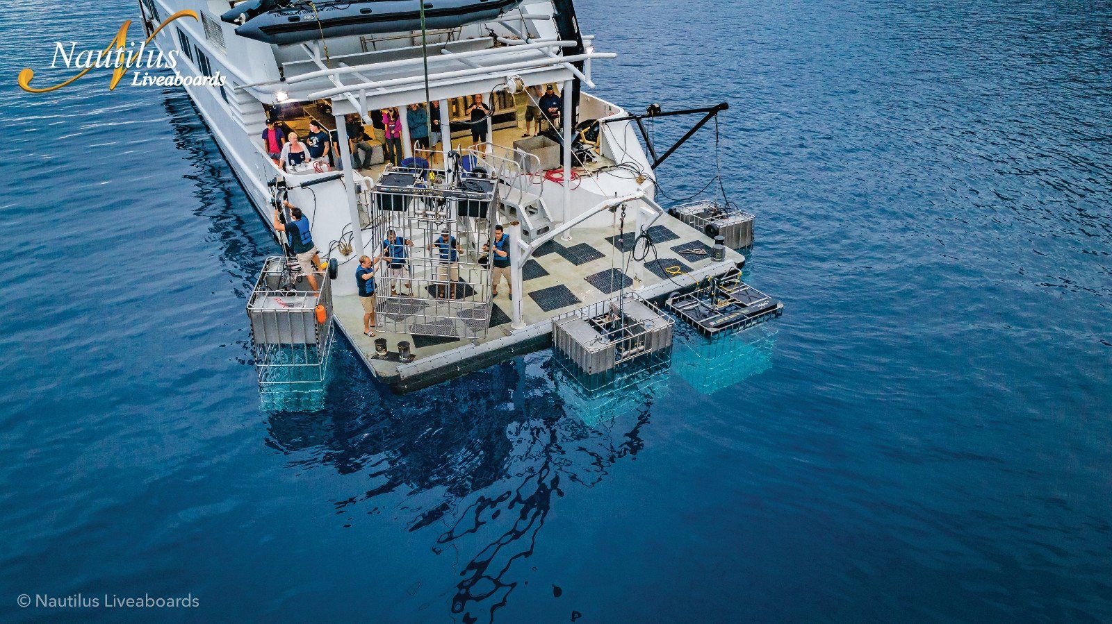 Large white boat in blue water with people on deck, gear platforms, and submerged viewing areas.