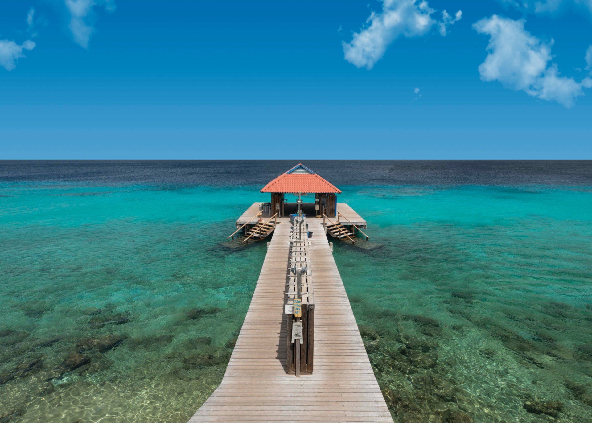 Wooden pier extending over clear turquoise water towards a small covered structure. Blue sky.