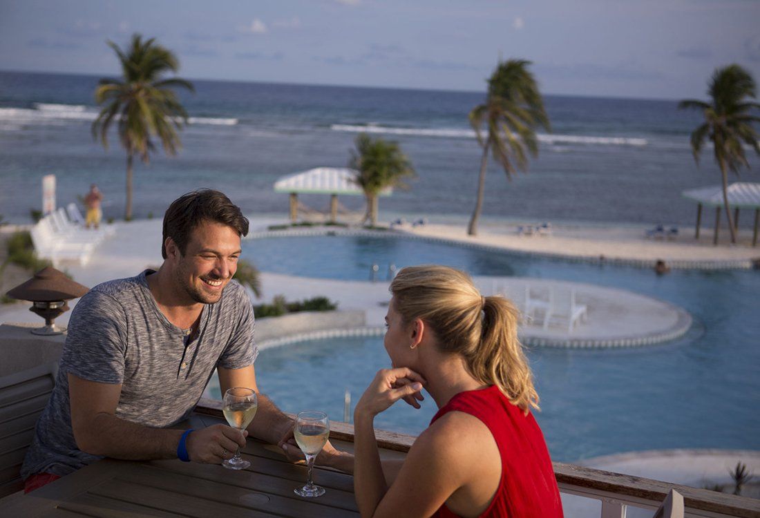 Couple smiling, drinking wine by a pool overlooking the ocean, palm trees in the background.